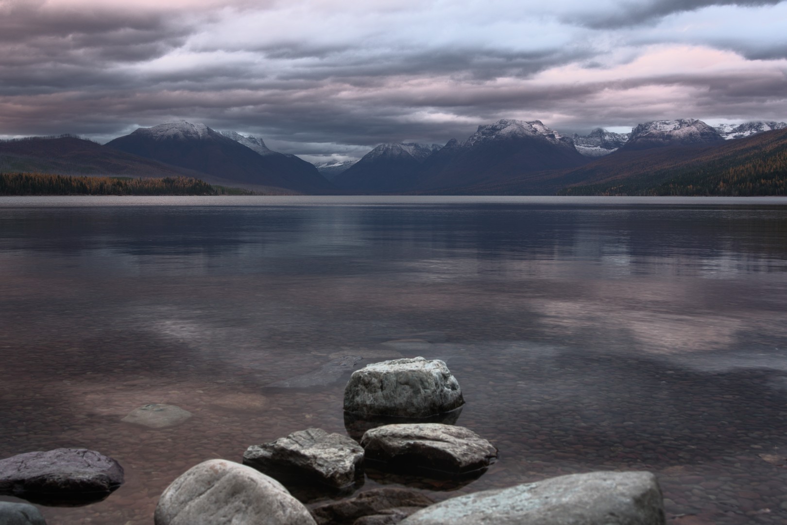 A serene still lake photo with glacial rocks in the foreground and mountains with snowy peaks in the background with sunset hue cloud cover.