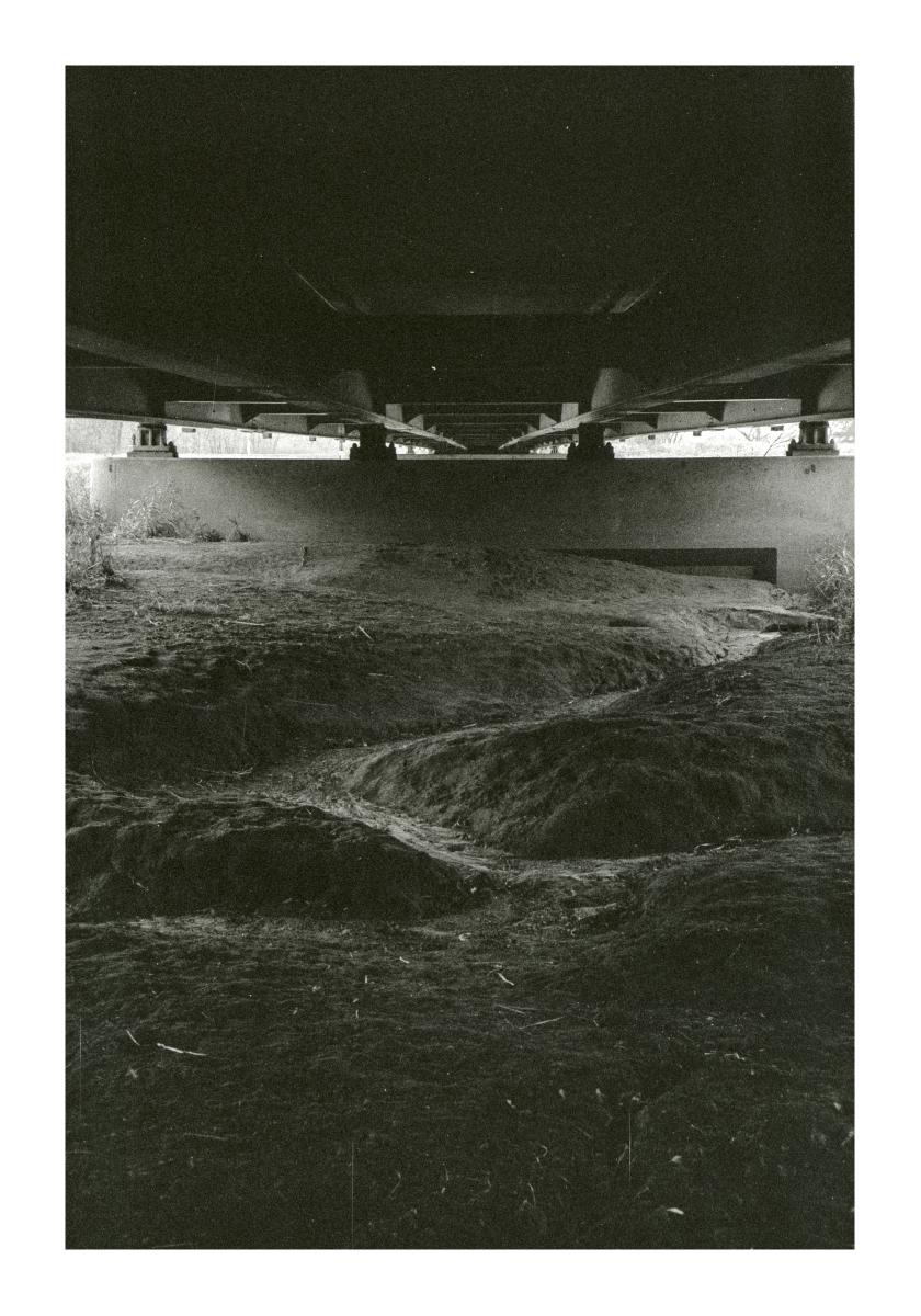 Black and white photo from under a roadway where the supporting steel and concrete form the ceiling and the dirt shaped by the flow of water is the floor.