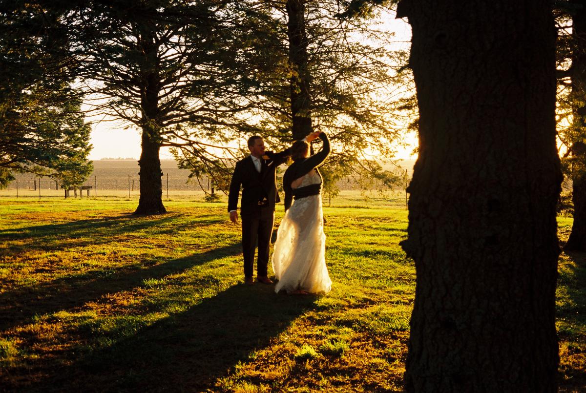A newly wed couple in a white dress and dark suit are slow dancing in a windbreak of connifer trees with warm golden hour light pouring in from the right and creating a glowing effect reflected in the expresson of joy.