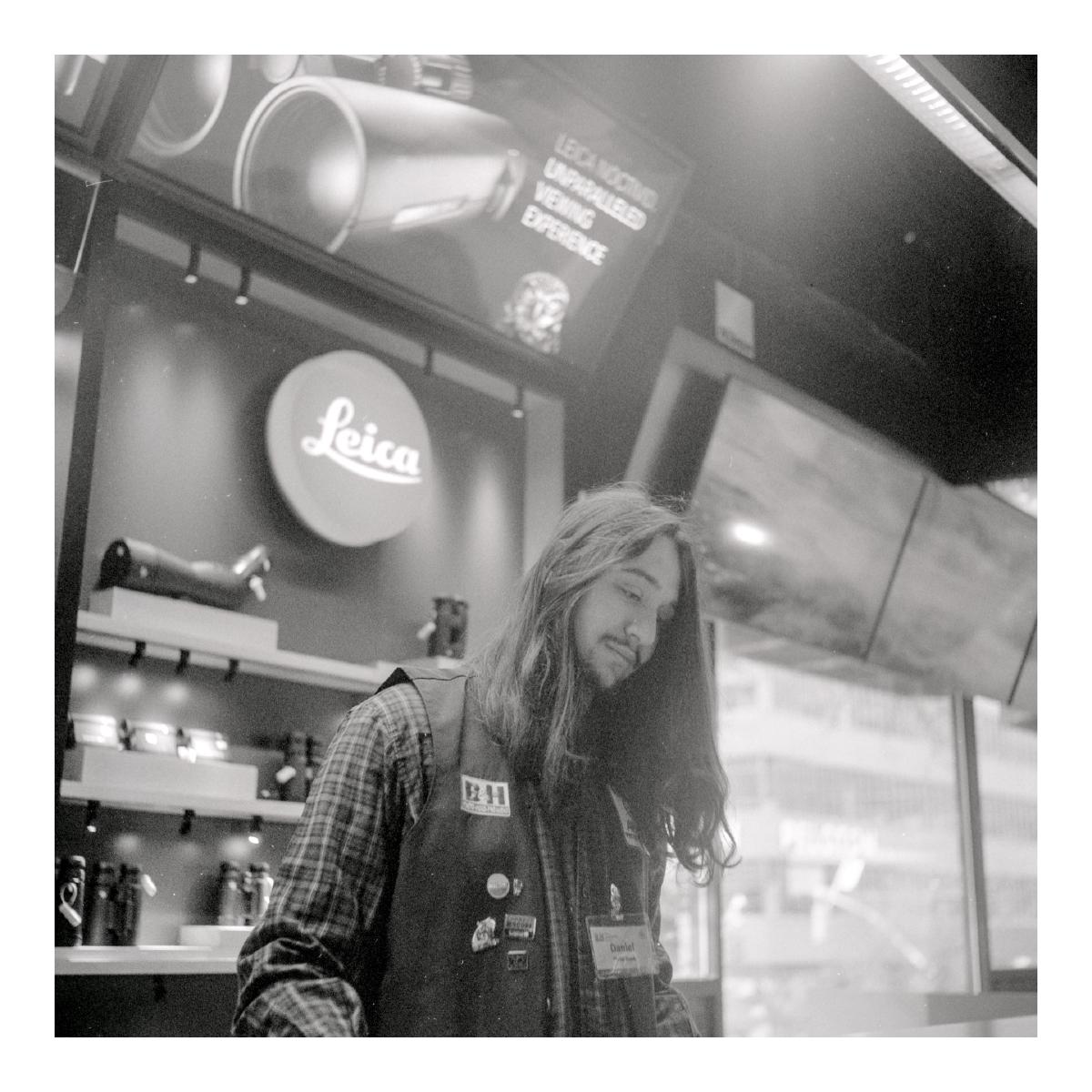 A person with long hair is looking down with a pleasant expression and standing at a counter. Behind the counter some binoculars and scopes are visible under a 'Leica' logo.