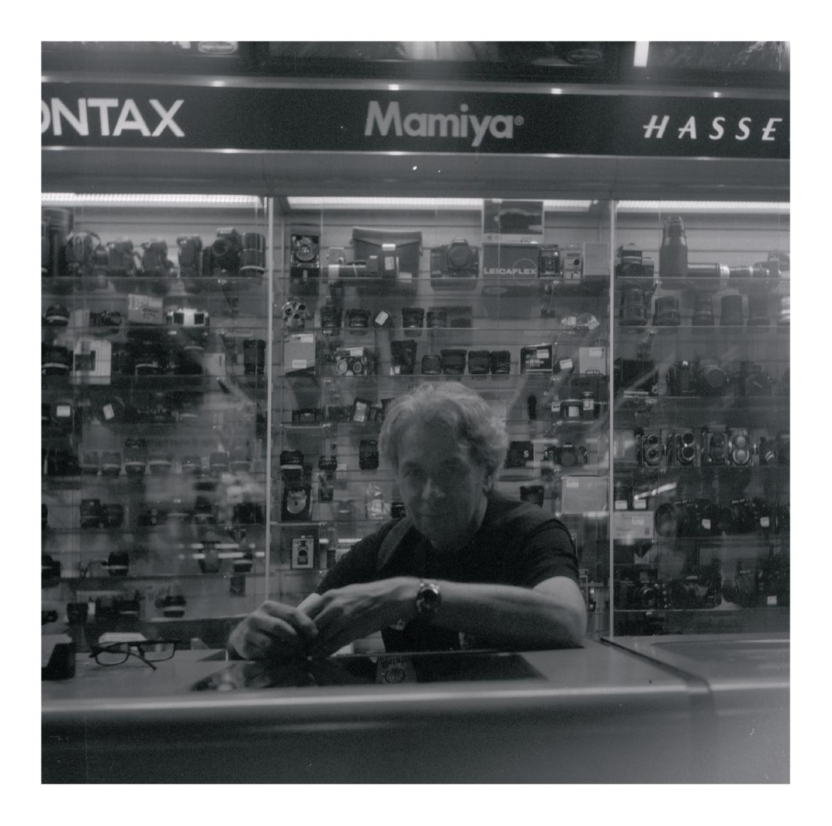 A person is sitting at a desk with their arms on the desk smiling at the camera. Behind them is shelves with rows and rows of film cameras. Some Brands are visible in the background Mamiya being the only one not cut off, Contax and Hasseblad logos are partially visible.