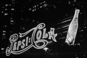 Black & White photo of a large neon sign at night time reading Pepsi:Cola in the Pepsi stylized sign with a large Pepsi glass bottle to the right of the sign also lit up. High-rise buildings' windows are the only things that can be seen directly behind the sign everything else is almost pitch black.