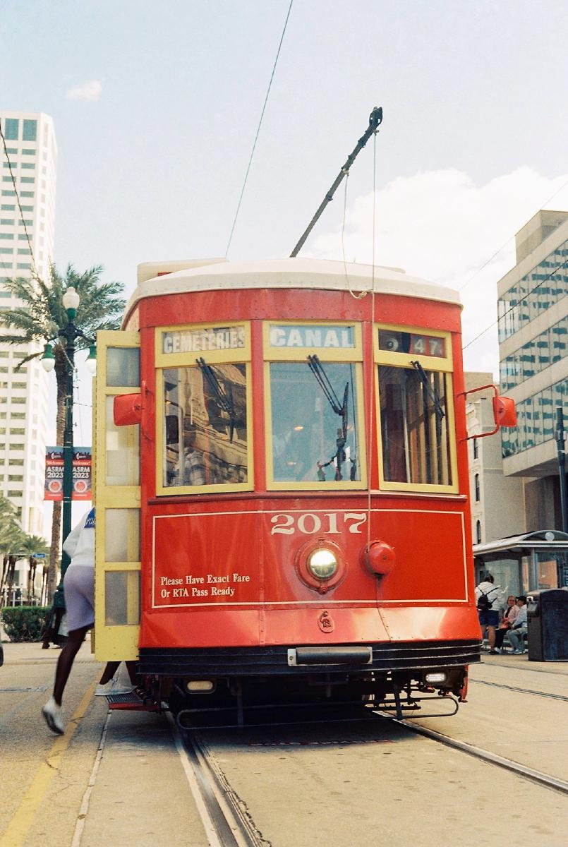 A red street car with yellow trim and doors with the number 2017 above a central round silver 'nose' right under a bay window style glass panes with the left pane reading 'CEMETERIES', center pane reading 'CANAL', and right pane showing '47'. A person is getting on the tram from the feint door, just one foot is seen leaving the ground. There are building and a palm tree in the background.