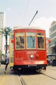 A red street car with yellow trim and doors with the number 2017 above a central round silver 'nose' right under a bay window style glass panes with the left pane reading 'CEMETERIES', center pane reading 'CANAL', and right pane showing '47'. A person is getting on the tram from the feint door, just one foot is seen leaving the ground. There are building and a palm tree in the background.