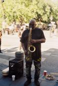 A street musician is standing in on a sidewalk and front of a cross walk with some people and trees which are in the bright sunshine. The musician standing in the shade is an older black man with short gray hair is playing a large shiny saxophone wearing a wearing t-shirt and black jeans with serval colorful patches. 