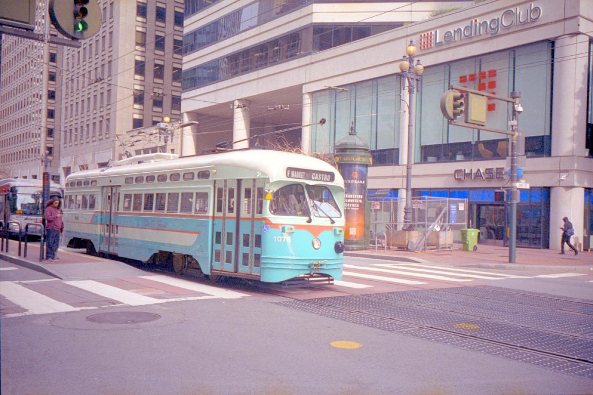 A San Francisco street car #107 from Market to Castro is white on top, baby blue on the bottom, and an orange stripe through the middle is stopped at an intersection. There are a few pedestrians on either side of the street looking at their phones.