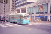 A San Francisco street car #107 from Market to Castro is white on top, baby blue on the bottom, and an orange stripe through the middle is stopped at an intersection. There are a few pedestrians on either side of the street looking at their phones.