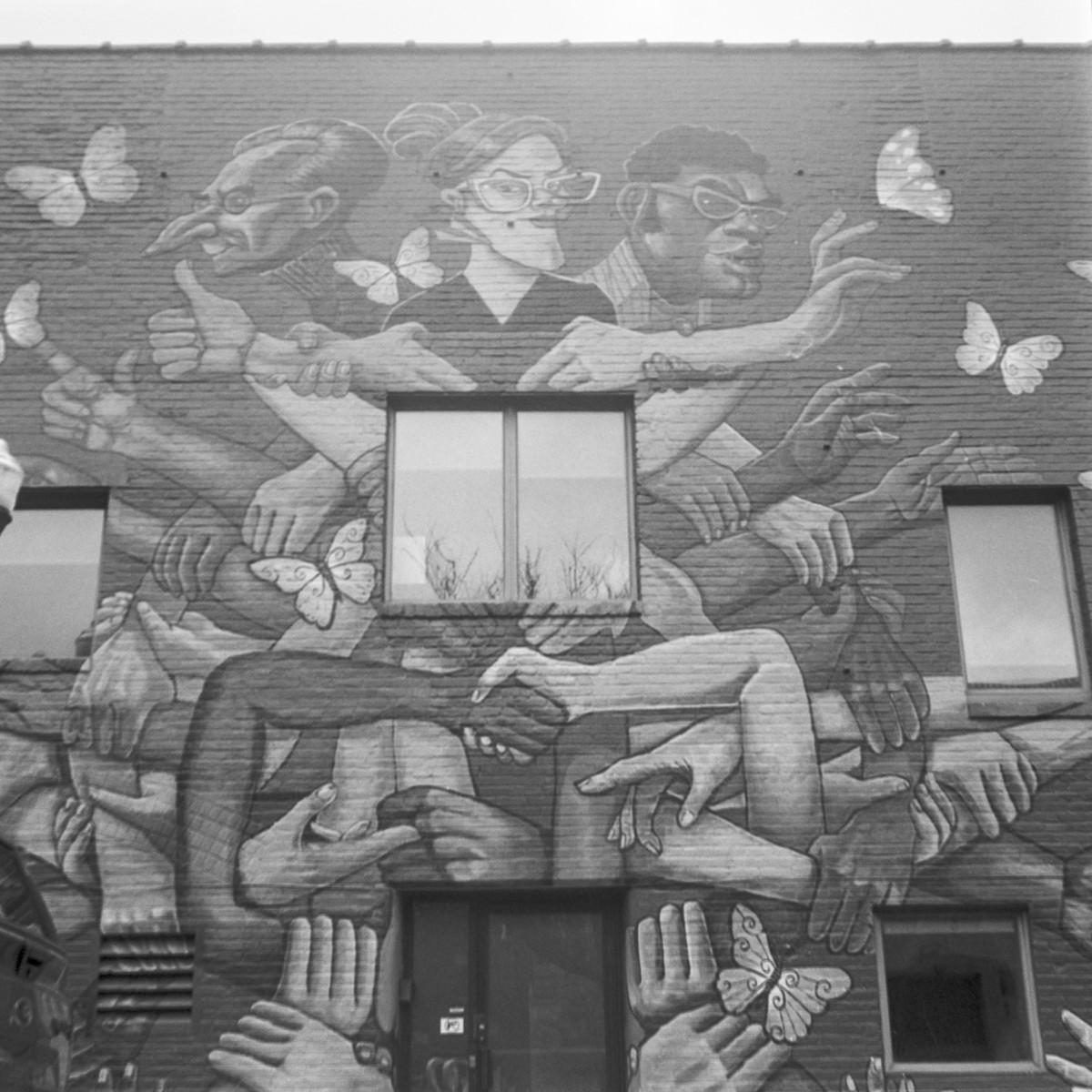 Black and white photo of a building facade with a few windows and a door at the bottom with a mural painted on the entire front. The mural has three people with distinct looks and many hands holding each other and some reaching out to butterflies.