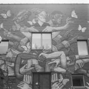 Black and white photo of a building facade with a few windows and a door at the bottom with a mural painted on the entire front. The mural has three people with distinct looks and many hands holding each other and some reaching out to butterflies.