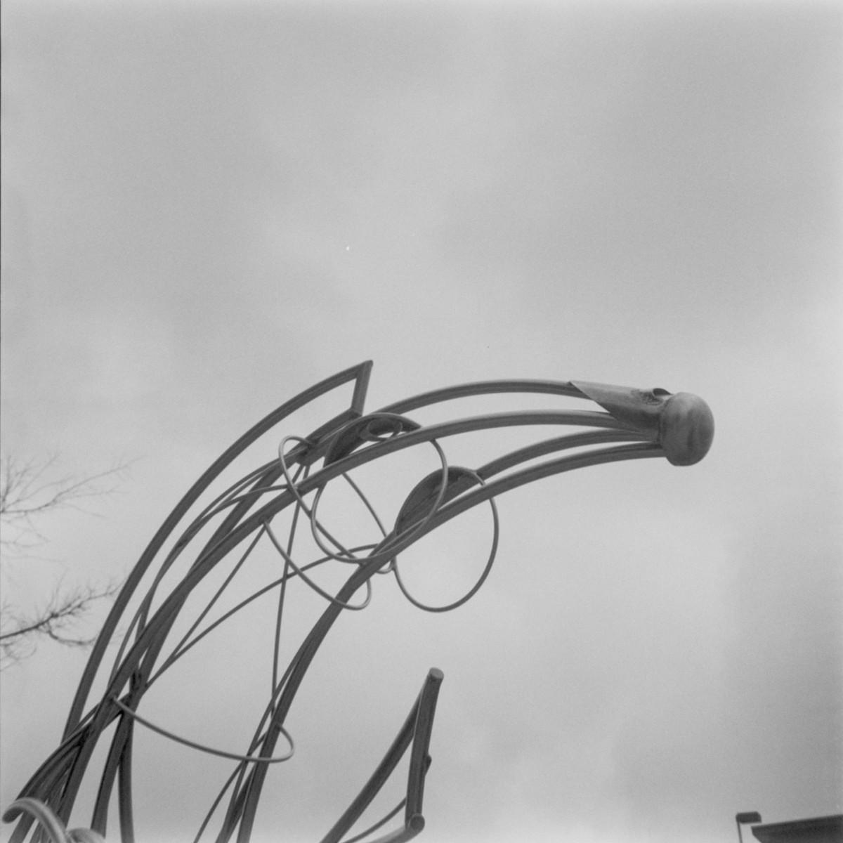 Black and white photo of a metal sculpture of an abstract bee stinger (open to interpretation) made of pipes and circles that are capped off in a bulb, against a stark and gray sky.