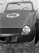 Black and white photo of an old convertible sports car with 66 painted on the hood inside a white circle, the back of the car is curiously missing but not immediately obvious.