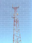 In camera double exposure: A radio tower made out of lattice construction and painted red and white with several dish and cylindrical antennas sticking out against a blue sky is super imposed on top of a brick background.