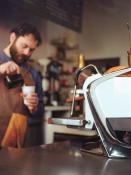 A high end commercial espresso machine is seen in profile in bright white and shiny chrome curves taking up a third of the frame right. As focus falls away to the mid-ground a barista wearing a grown apron, blue shirt, and a thick beard can be seen pouring milk from a pitcher into a cup. The background almost totally blurry is coffee shop standards like tall grinders, shelves with brewing tools.