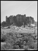 Black and white photo of a desert scene with shrubs and a few cacti in the fore and midground in front of a massive rock outcrop with jagged peaks and vertical cliff faces.