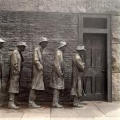 Color photo of life size bronze sculptures in front of a black brick facade with a black door on the right end of the frame. The five figures are facing towards the door, they're all of different heights but dressed similarly in long trench coats and slight variations of a trilby style hats. The expressions of the statues are gaunt, most have their hands in their coat pockets the one in the front of the line has his arms crossed. The bronze statues have the greenish patina except for the shoulders and sleeves where people have touched them, almost as if in support for their plight as if lending strength to them to stay standing in the bread line despite the inclement times and weather. The writing above is mostly cutoff except for the word 'NOURISH'.