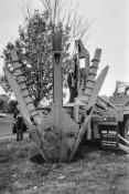 A massive tree planting mechanized spade is on the back of a truck with a tree loaded in the spade. The spade is being lowered with the tree into a hole in the ground. The mudflap on the truck says 'BIG JOHN Manufacturing'.