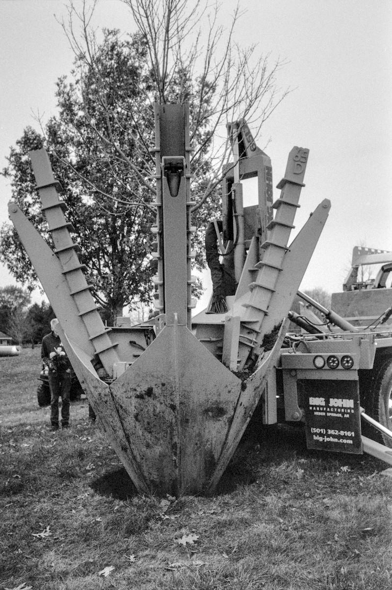 A massive tree planting mechanized spade is on the back of a truck with a tree loaded in the spade. The spade is being lowered with the tree into a hole in the ground. The mudflap on the truck says 'BIG JOHN Manufacturing'.