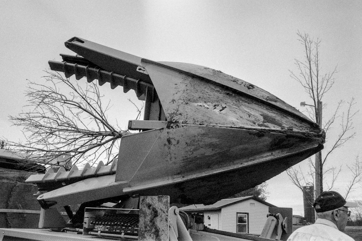 A massive tree planting mechanized spade is on the back of a truck with a tree loaded in the spade. There is a person's head and shoulder wearing a baseball cap in the bottom right, which clues in the scale of the spade to be about 10ft tall from tip of the blade to the top of the pivot arm and about the 15 ft in diameter.