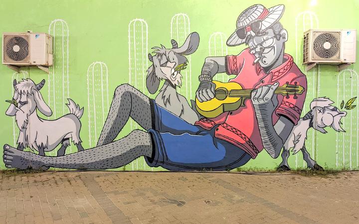 Street mural of a man wearing a straw hat, red shirt, blue shorts, reclining while playing a ukuele while three goats surround him chewing leaves with funky expressions.
