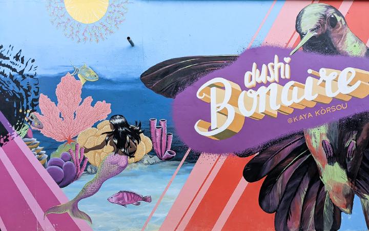 Wall mural of an underwater scene with corals and fish with a mermaid floating and taking the scene in. On the right side there's a layer of stripes with a massive hummingbird seeming to hold up a sign reading 'dushi Bonaire @kaya korsou'