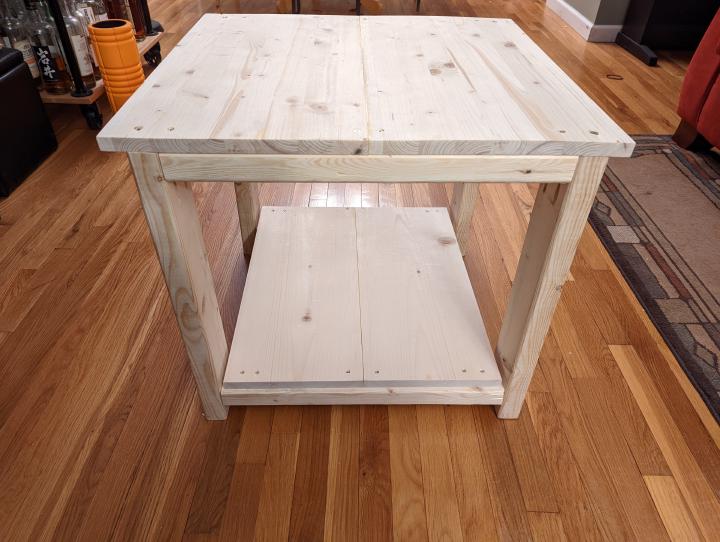 A small table made of light colored pinewood construction lumber with a table top and a bottom shelf made from similar colored thin boards. You can see several fastening screws and not everything is quite plumb but functional