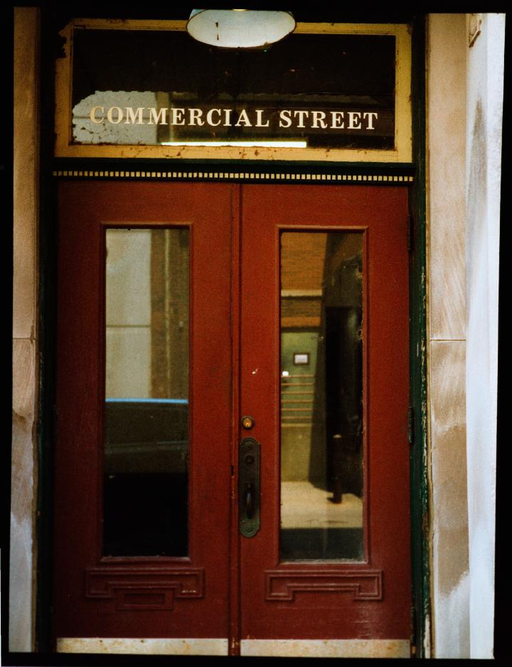 Color of a red door with two panes each with a glass window. Above the door is is another horizontal pane of glass with the words 'COMMERCIAL STREET' emblazoned in gold. The door has trim decoration a heavily patina'd copper door handle and some shiny bronze splash guards at the bottom. The opposite side of the street is reflecting a nondescript scene of an alley, the reflection of the photographer is missing. The vibe is of by gone glory fallen into disrepair.