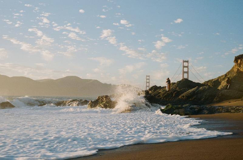 A sunset scene at Baker Beach by the Golden Gate Bridge in San Fransisco, CA, USA. The sun is setting off screen to the left and casting a beautiful light sideways towards the subjects which is further scattered by the spray in the air. A wave is crashing in from the left with white foamy water rushing onto the sand and covering most of the bottom third, beyond which are the rocks that have caused a huge splash of brilliant white spray just in front of a couple that is standing front to back on some rocks hugging framed in between to two towers of the Golden Gate Bridge in the background which stands under a blue sky with scattered fluffy clouds. Shot with a Voigtlander BessaR3M and Voigtlander40f1.4MC lens, on KodakKodacolor100, and developed with LabDev by Shom Bandopadhaya. Licensed under Creative Commons Attribution-NonCommercial-ShareAlike (CC BY-NC-SA).
