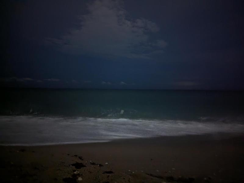 A dark image of the ocean at night but the white foam from the crashing wasves on the beach is visible against the greenish blue of the water and the dark blue of the sky with some gray storm clouds and a hint of lightning in the distance.